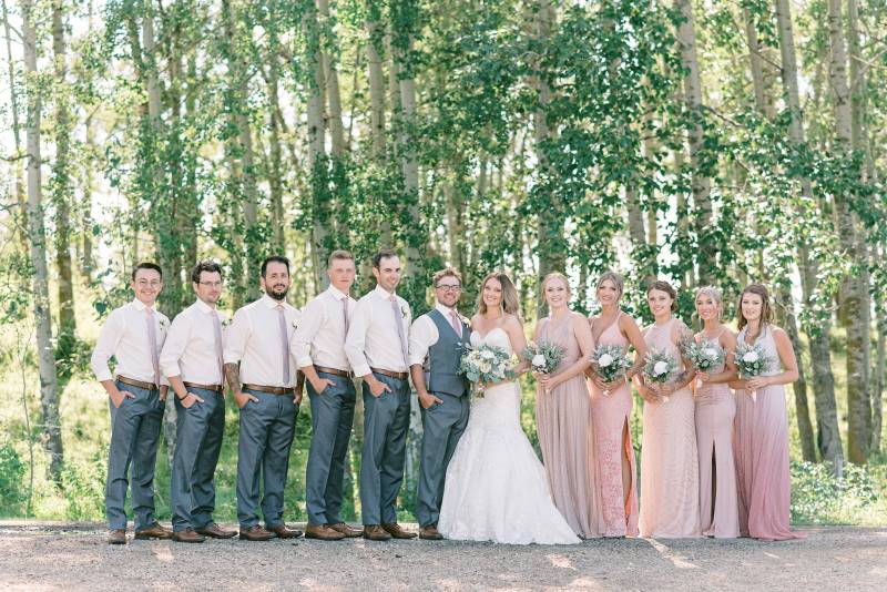 Bride and groom stand together beside bridesmaids and groomsmen on gravel path 