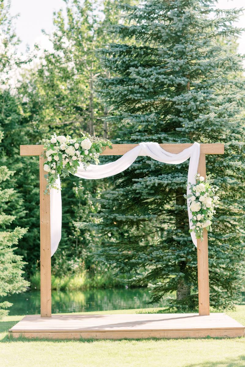 Wooden wedding arch on wooden platform with handing white fabric and white floral arrangements