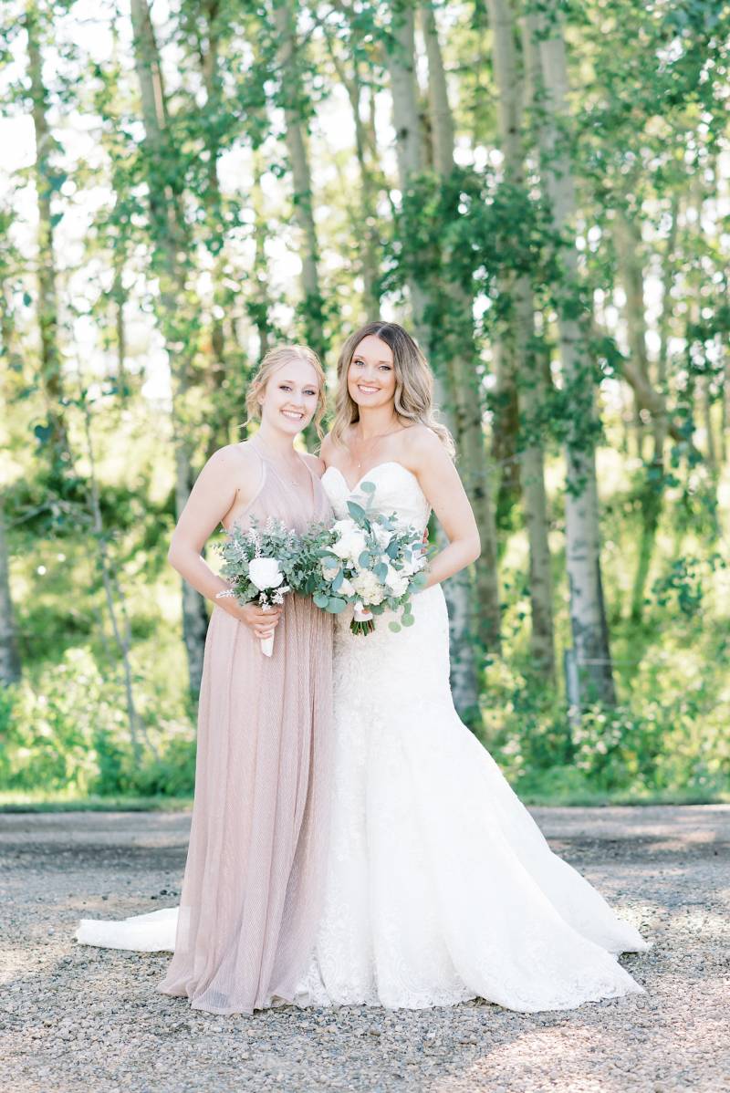 Bride and bridesmaid stand together smiling holding white bouquet 
