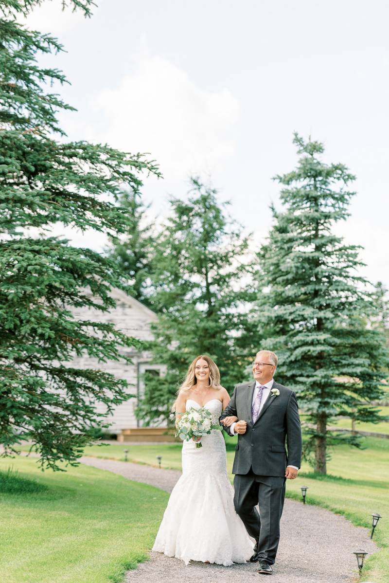 Bride in white dress holding bouquet and father walk down dirt pathway 